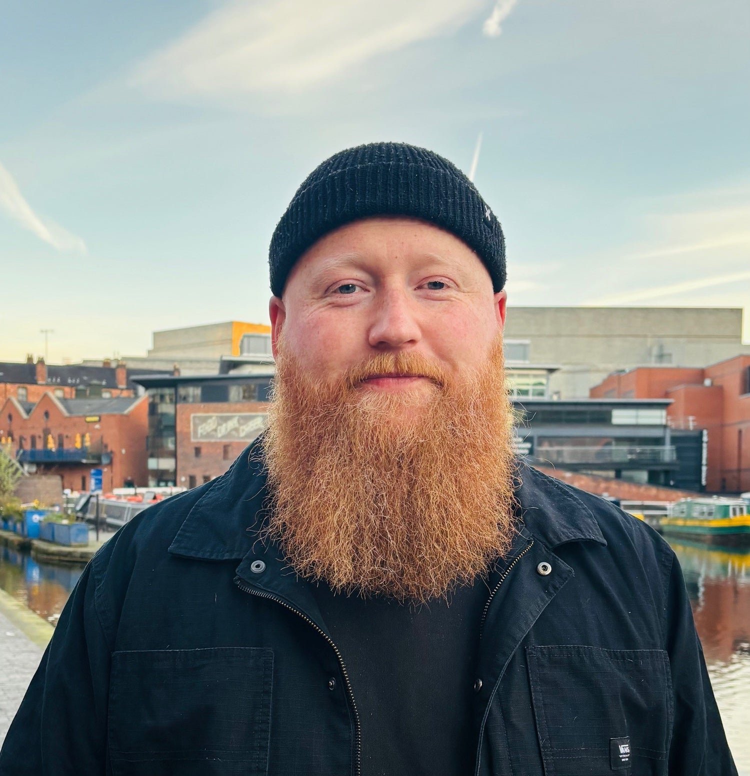 Portrait of a Jack the home enable founder with a long red beard wearing a black beanie and jacket, standing by a canal with modern and historic Birmingham buildings in the background, looking warmly at the camera.